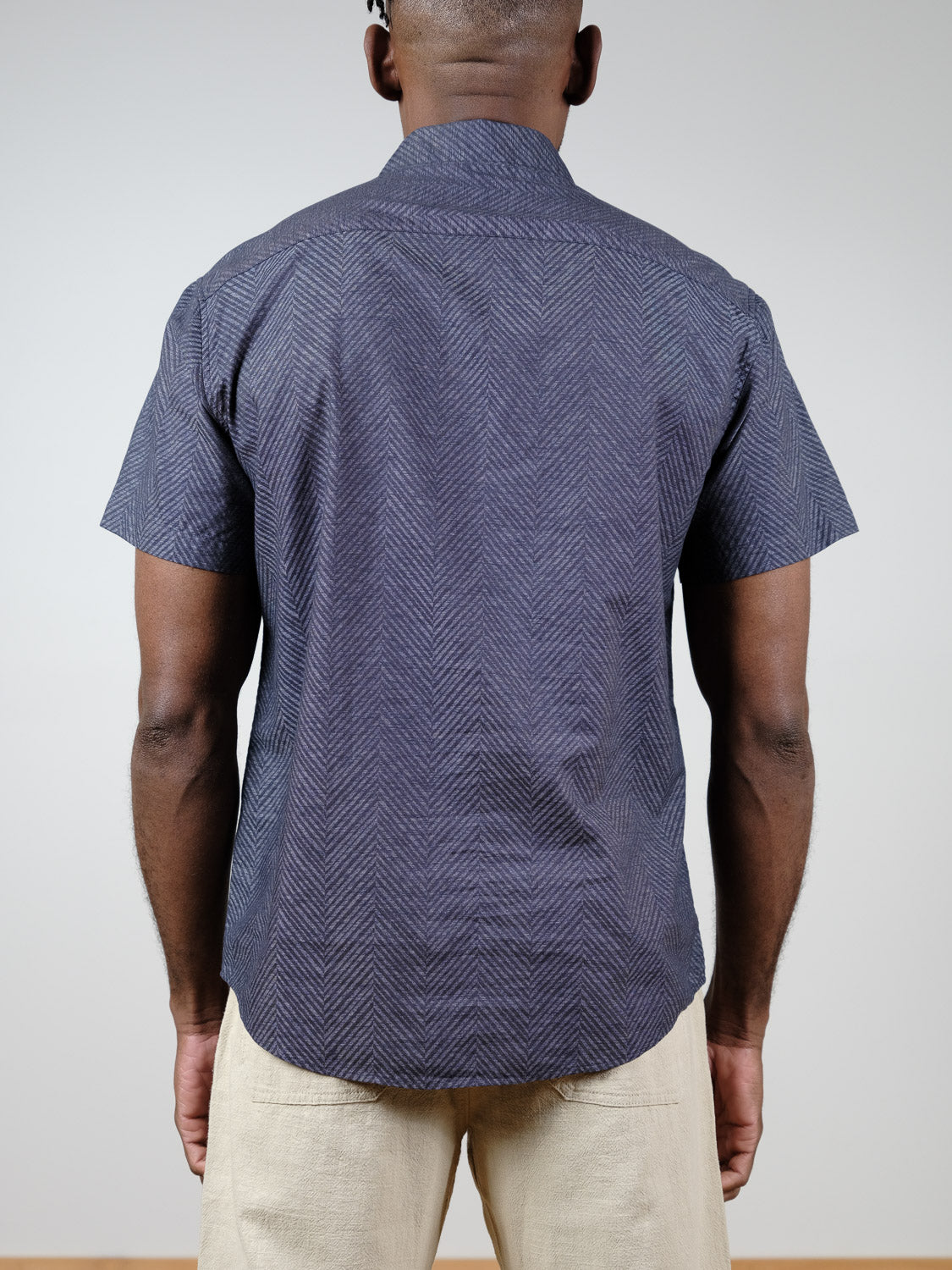 A man, with his back to the camera, wears the Reveller Short Sleeve Shirt by ID Menswear—a relaxed-fit dark geometric weave button-up—paired with light beige pants against a plain background.