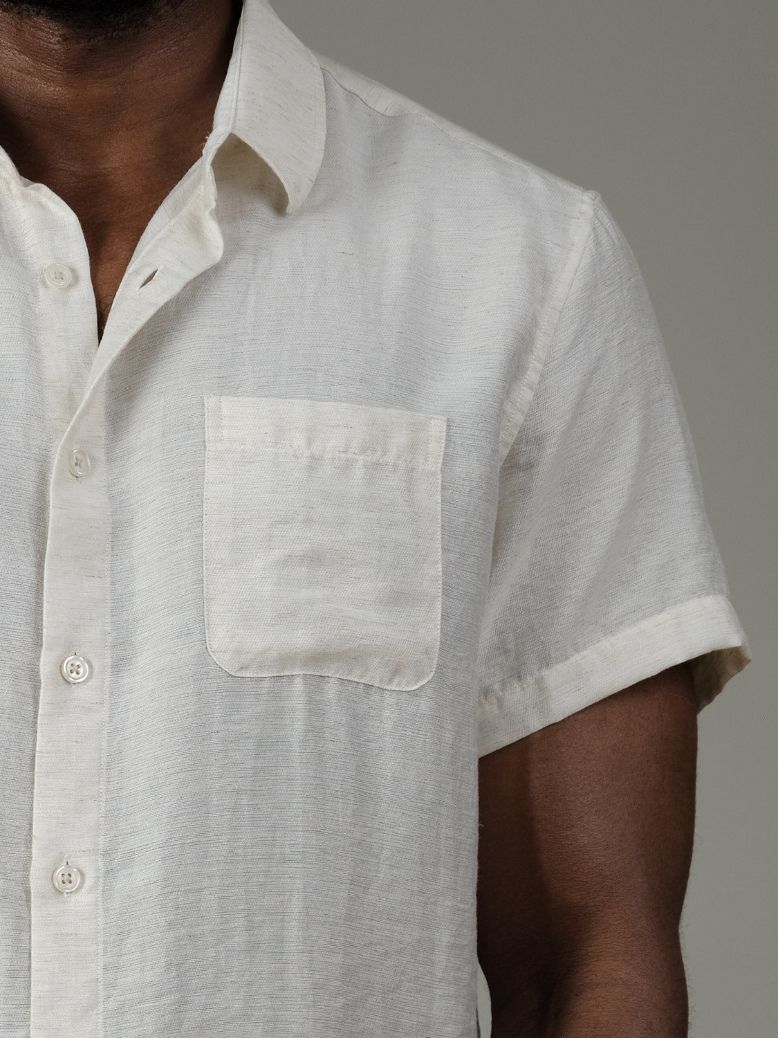 A person wears the Hiker Short Sleeve Shirt by ID Menswear—an off-white linen button-up with a chest pocket—shown from shoulders to mid-upper arm against a plain background, making it an ideal pick for summer basics.