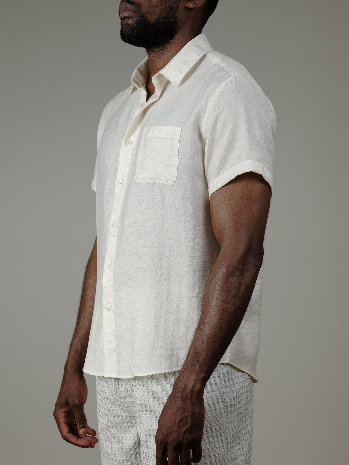 A man wearing the ID Menswear Hiker Short Sleeve Shirt in cream—a relaxed fit linen button-up with a chest pocket—paired with light textured shorts, stands against a plain gray background.
