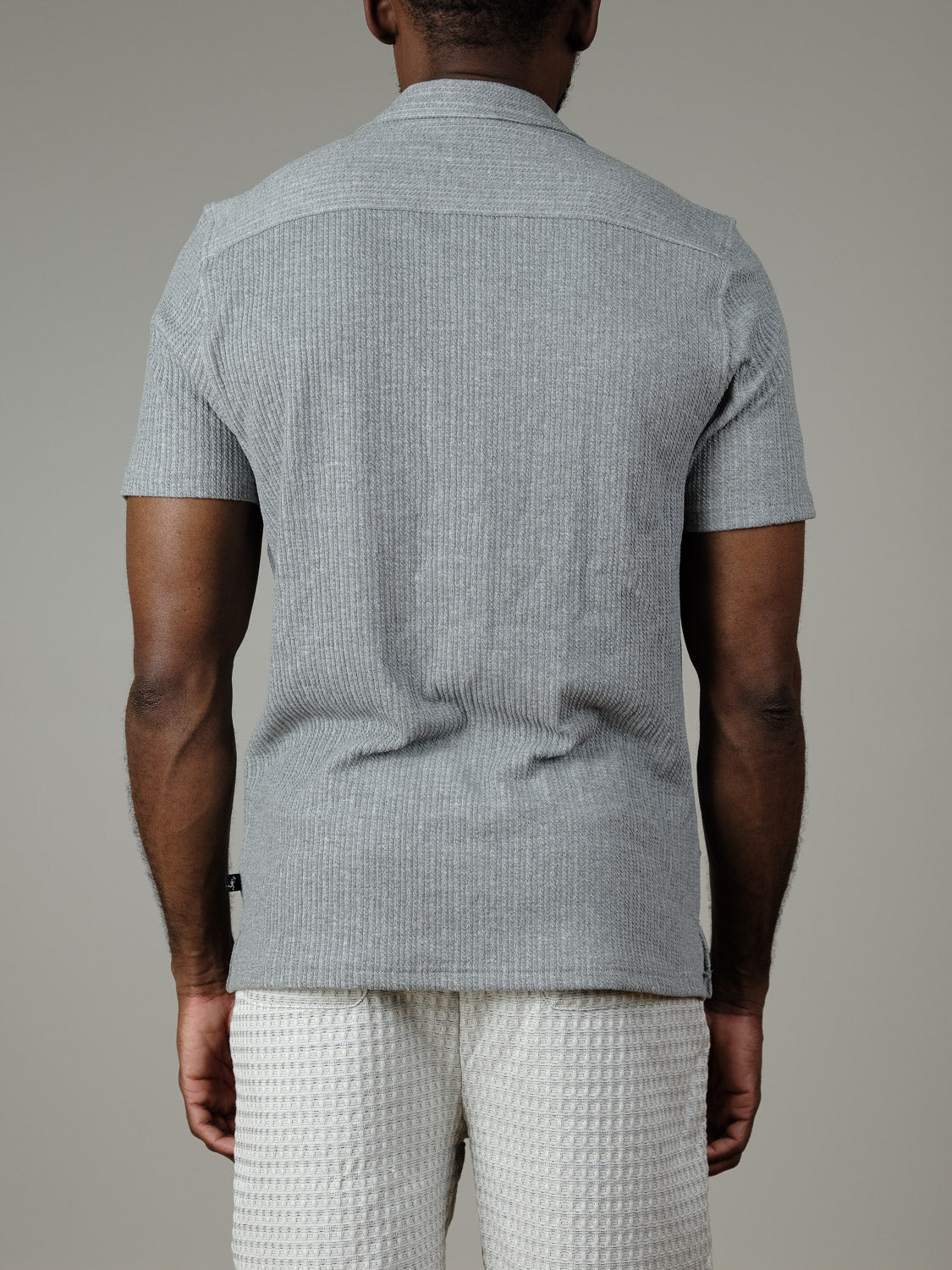 A person stands facing away from the camera, wearing the Civil Society Viggo Knit Resort Shirt in light gray, paired with off-white waffle-patterned shorts, set against a plain, light gray background.