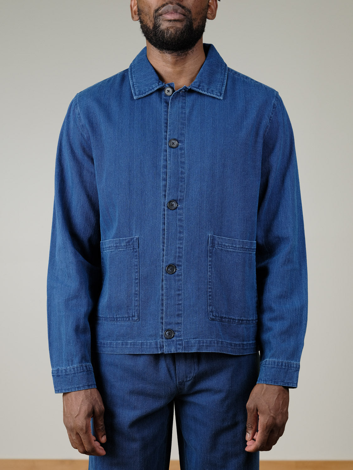 A man dressed in the Casual Friday Asplund Herringbone Jacket, featuring front pockets and a blue hue with matching pants, stands against a plain light background. His face is partially out of frame.