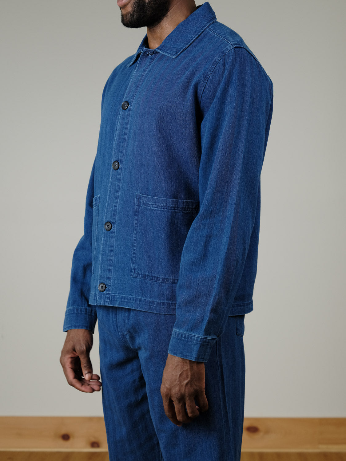 A man models the Casual Friday Asplund Herringbone Jacket in blue, standing indoors against a plain background. The photo shows his upper body and lower face as he faces slightly to the side.