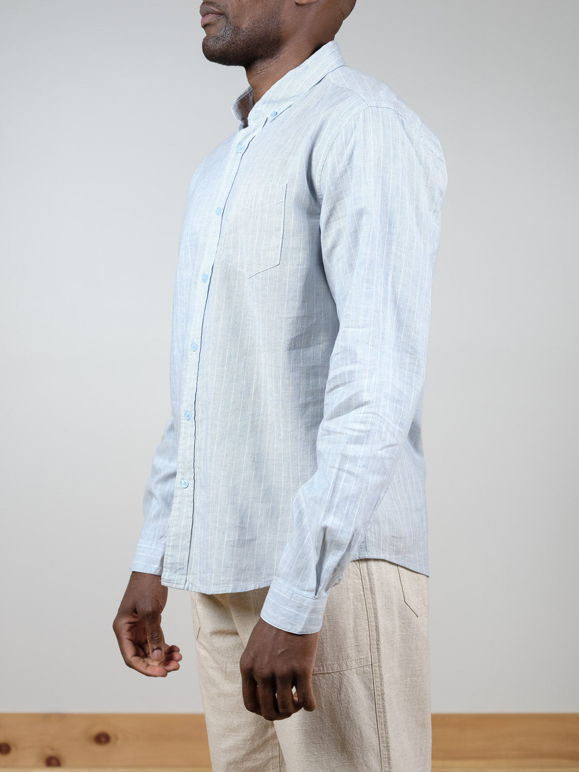 A man stands in profile indoors wearing the ID Menswear Dubai Long Sleeve Shirt, a light blue pinstripe shirt, paired with beige pants.