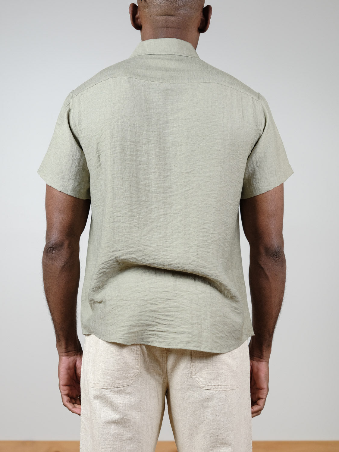 A person stands facing away, wearing the Hiker Short Sleeve Shirt in light olive by ID Menswear and cream-colored pants—a relaxed, classic summer look against a plain background.