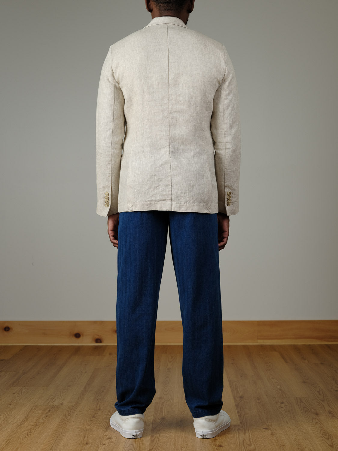 A person stands indoors on a wooden floor, facing away from the camera, wearing the Casual Friday Torup Linen Blazer in light beige, blue trousers, and white sneakers against a plain gray wall.