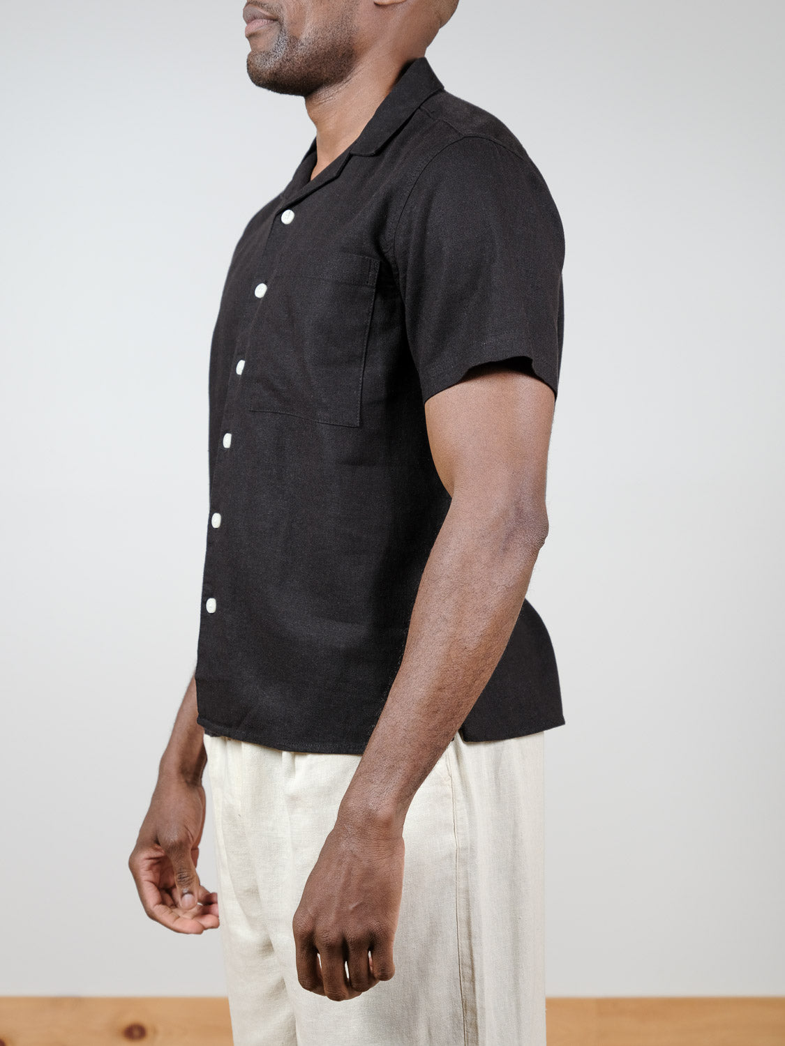 A person stands sideways wearing the !Solid Allan Cuba Woven Short Sleeve Shirt in black, paired with light beige pants against a plain background.