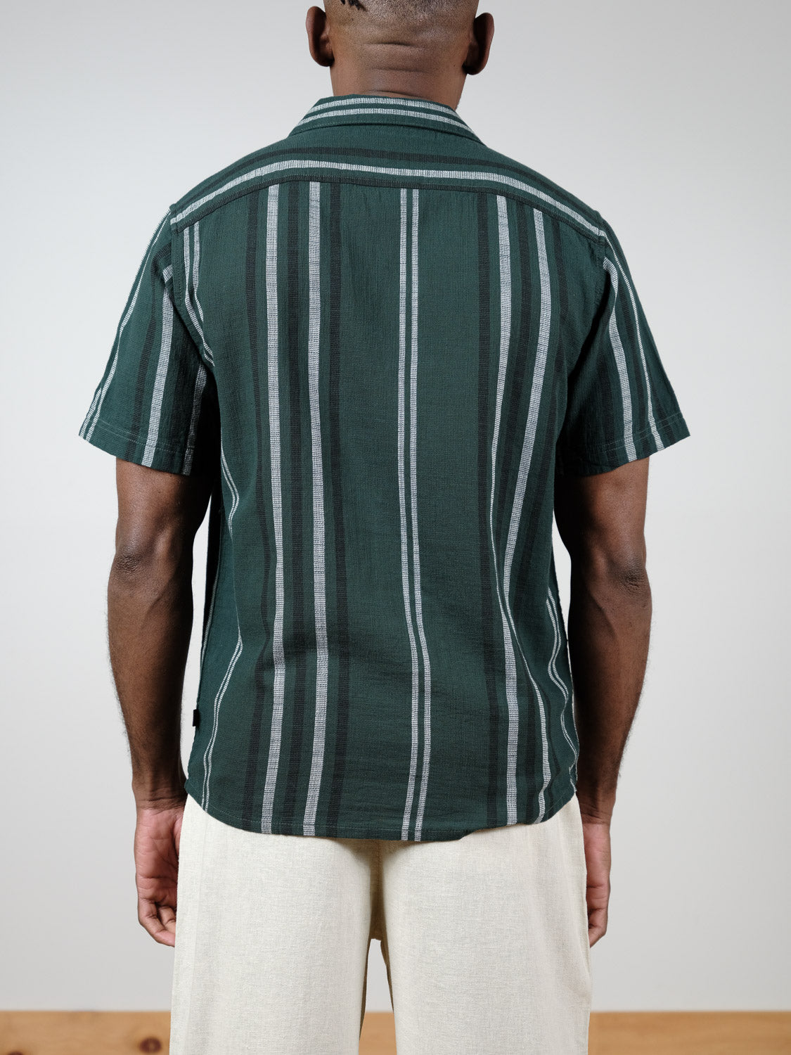 A person stands facing away, wearing the !Solid Isai Short Sleeve Shirt in green with vertical white and gray stripes, paired with light beige pants, against a plain light background.