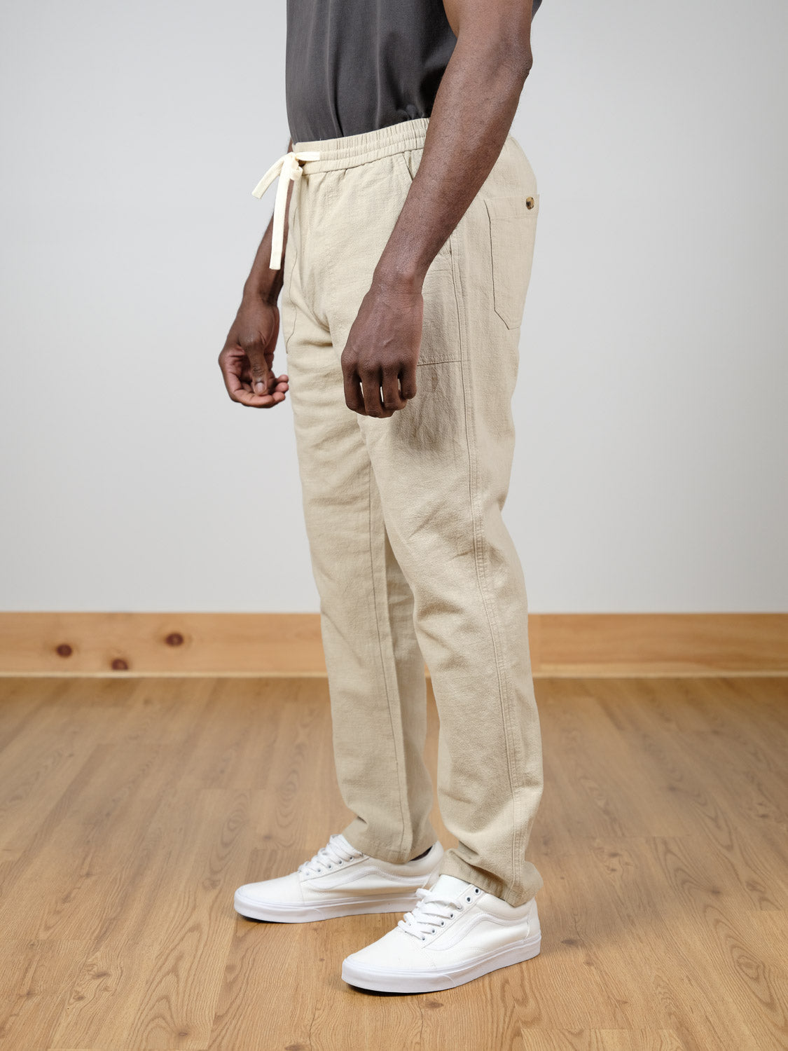 A person stands indoors on a wooden floor, wearing the beige Sahara Linen Cotton Pant by ID Menswear, paired with a dark gray shirt and white sneakers. The photo shows their body from shoulders down, arms relaxed at their sides.