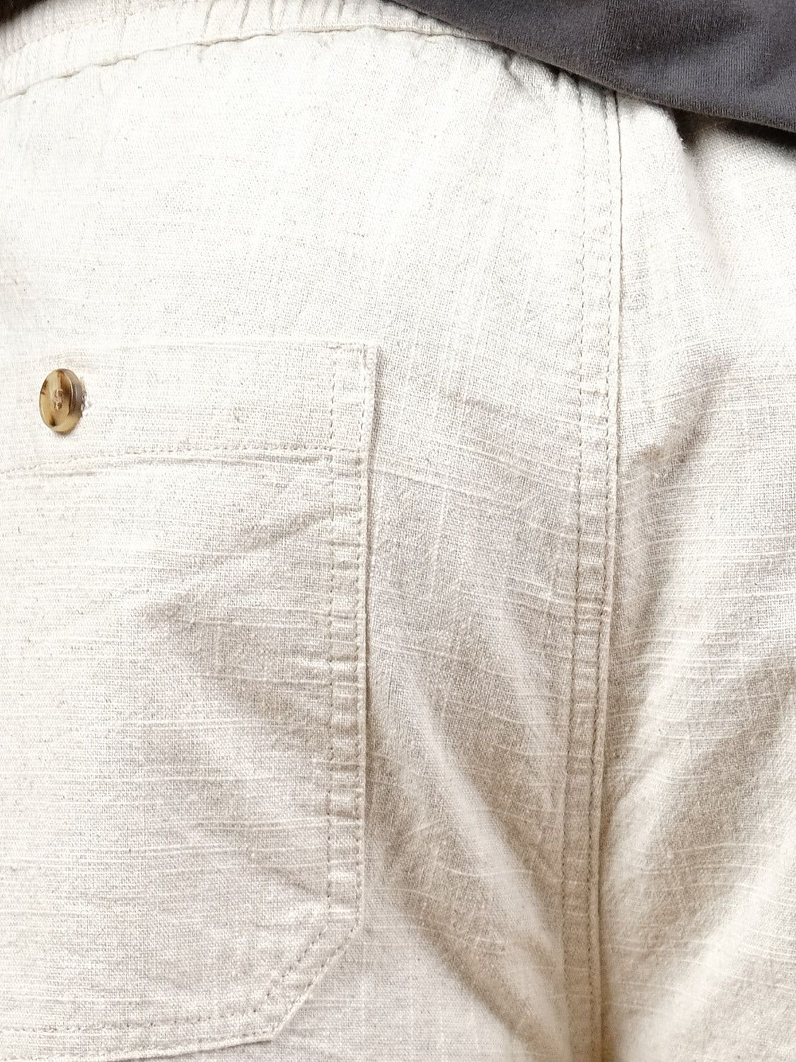 Close-up of the Sahara Linen Cotton Pant by ID Menswear in cream, featuring a relaxed fit, back pocket with a brown button, and part of a dark shirt at the waistband. The fabric has a natural, textured look.