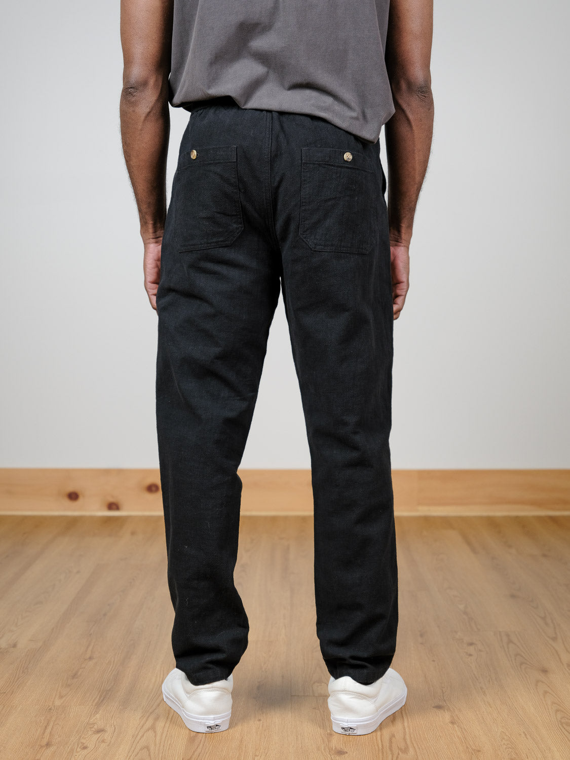 A person wearing a gray t-shirt, white sneakers, and black Sahara Linen Cotton Pant by ID Menswear stands on a light wood floor facing away from the camera.