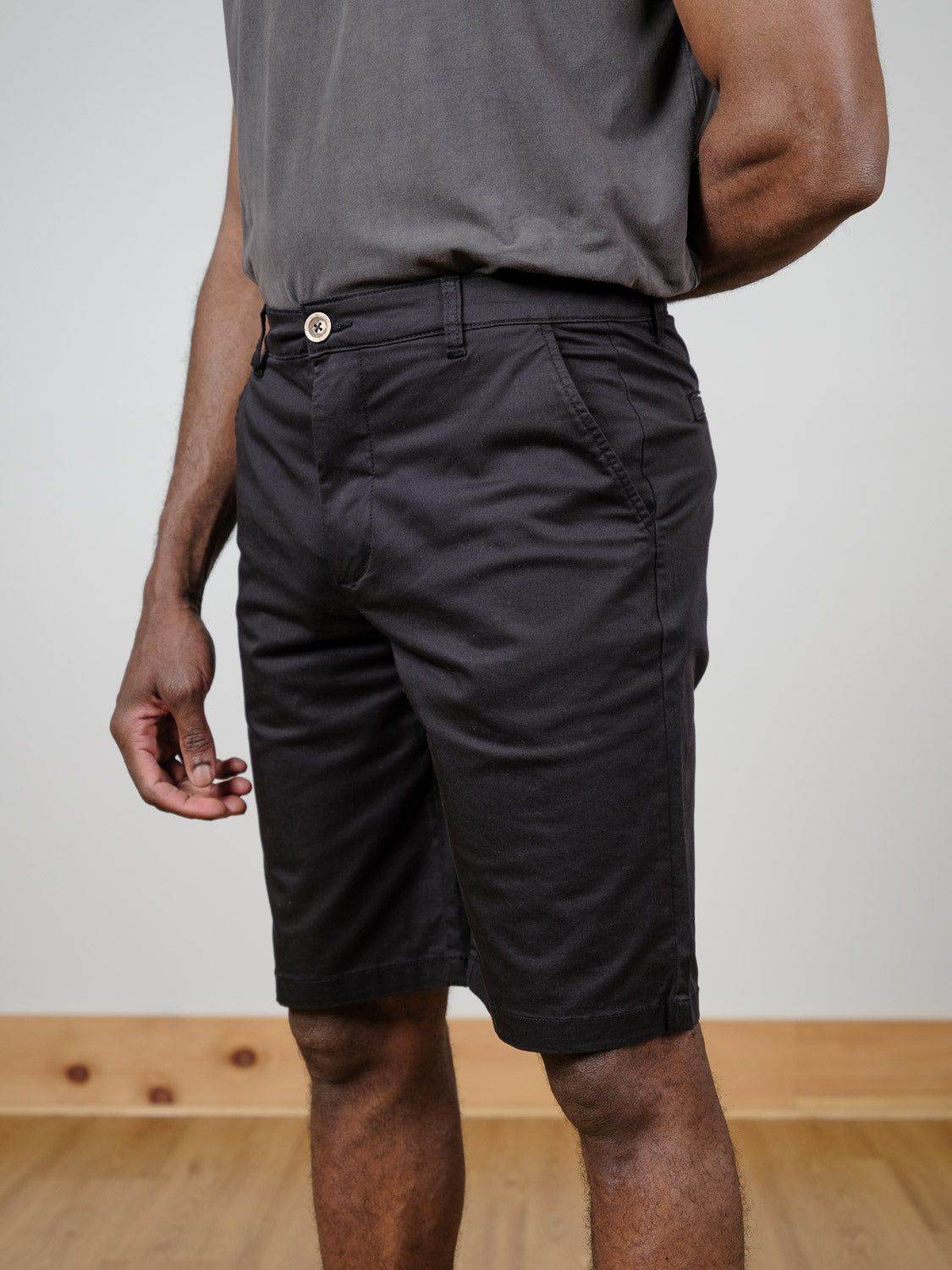 A person stands indoors on a wooden floor, arms at their sides, wearing a charcoal gray t-shirt and the !Solid Bishop Short in black. Photo is cropped at the shoulders.