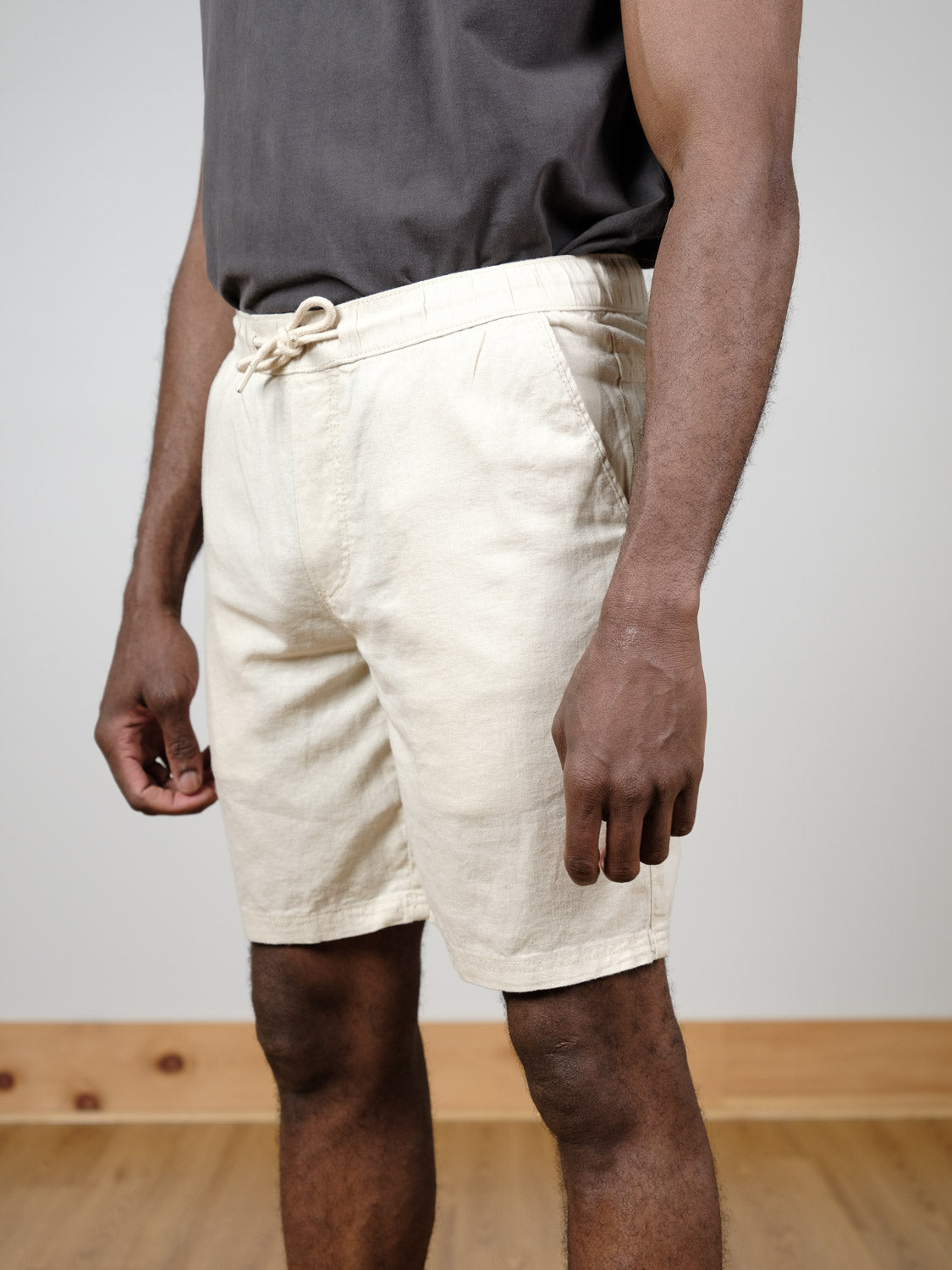 A person wearing a dark gray t-shirt and the !Solid Aurelius Elas Short in light beige stands indoors on a wooden floor, with a plain light-colored wall as the background.