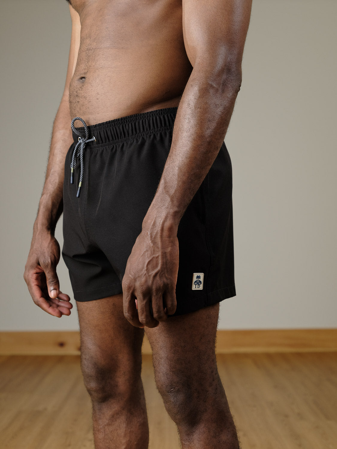 A man stands indoors wearing ID Menswear's Oyogu Quick Dry 4 Way Stretch Swim Trunk—black with a white patch logo on the left leg—showing his bare torso and legs against a wooden floor and light-colored wall background.