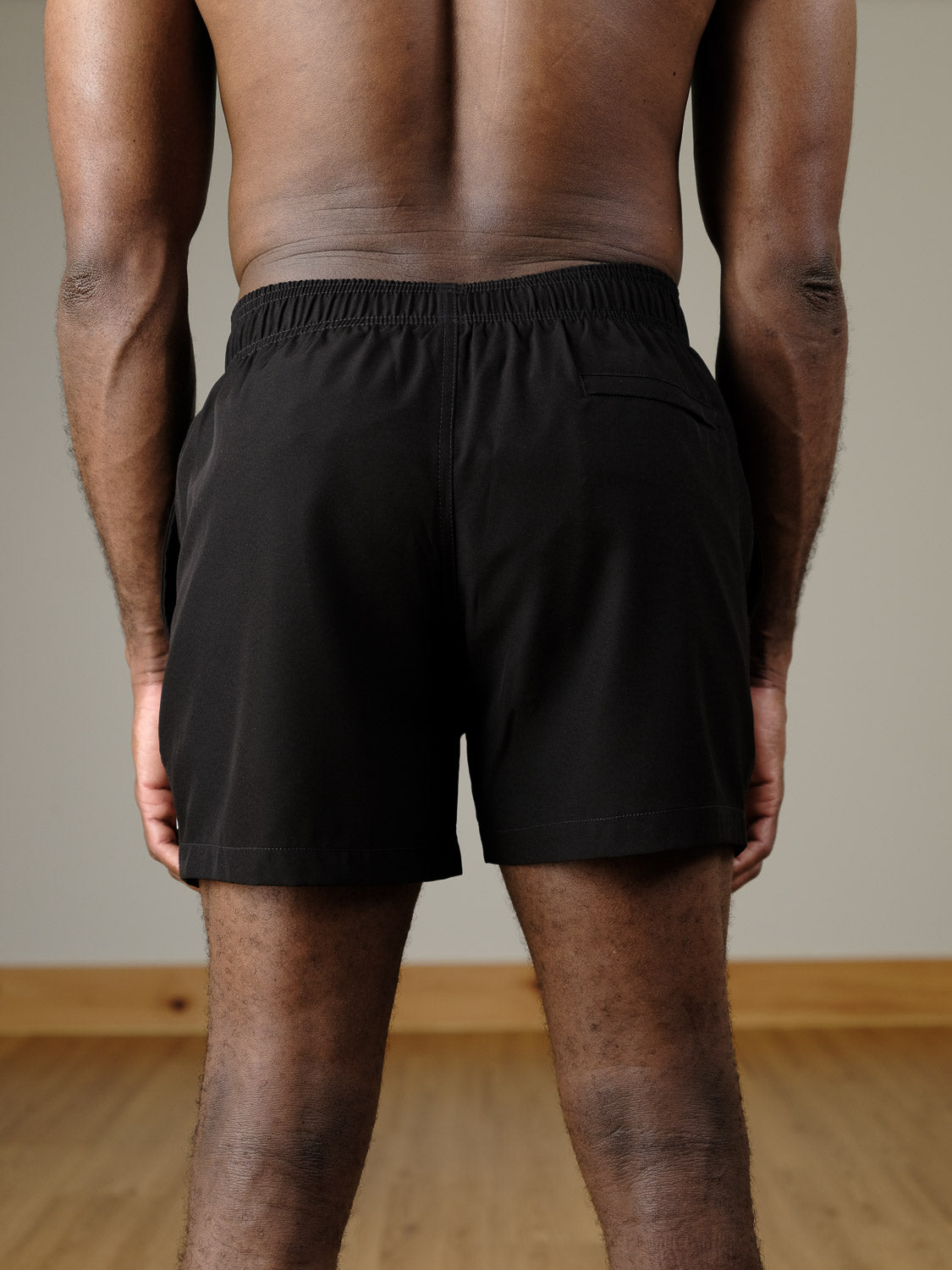 A person stands indoors with their back to the camera, wearing ID Menswear's Oyogu Quick Dry 4 Way Stretch Swim Trunk. The setting includes a wooden floor and a plain beige wall.