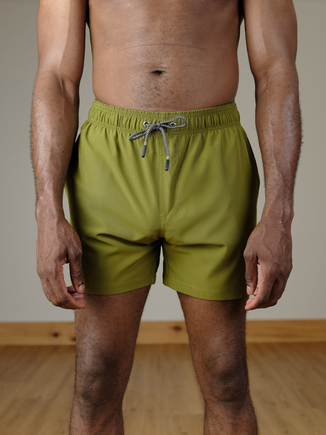 A man stands indoors from shoulders to knees, wearing ID Menswear's Oyogu Quick Dry 4 Way Stretch Swim Trunk in olive green. The setting includes a wooden floor and a plain beige wall.