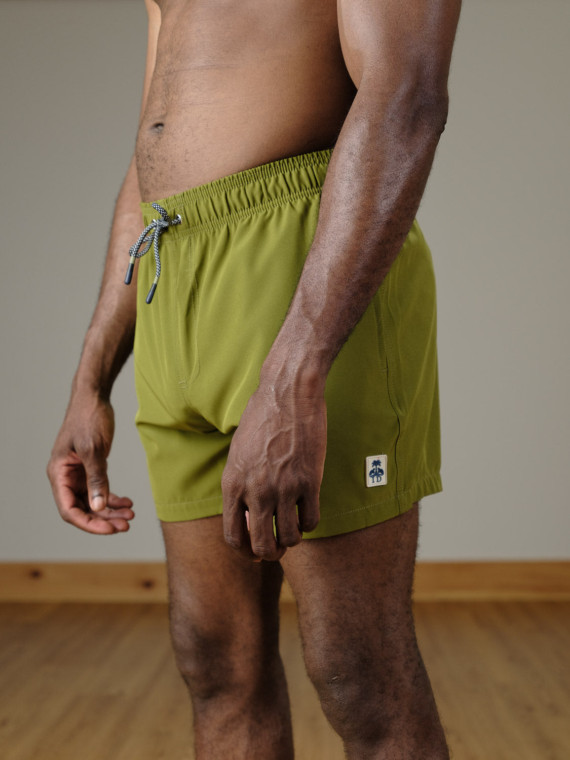 A person wears ID Menswear's Oyogu Quick Dry 4 Way Stretch Swim Trunk in olive green, standing indoors on a wooden floor, shown from mid-torso to mid-thigh.