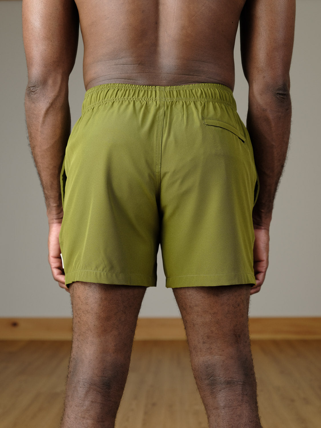 A person stands indoors with their back to the camera, wearing ID Menswear's Oyogu Quick Dry 4 Way Stretch Swim Trunk in olive-green, featuring an elastic waistband and back pocket. The setting is a plain wall and wooden floor.