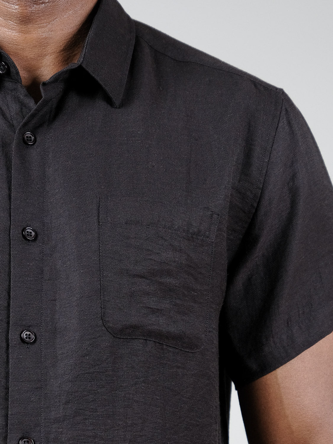Close-up of a person wearing the Hiker Short Sleeve Shirt by ID Menswear—a relaxed-fit, black button-up with a front chest pocket—shown from shoulders to mid-torso against a plain background. Perfect for summer basics.