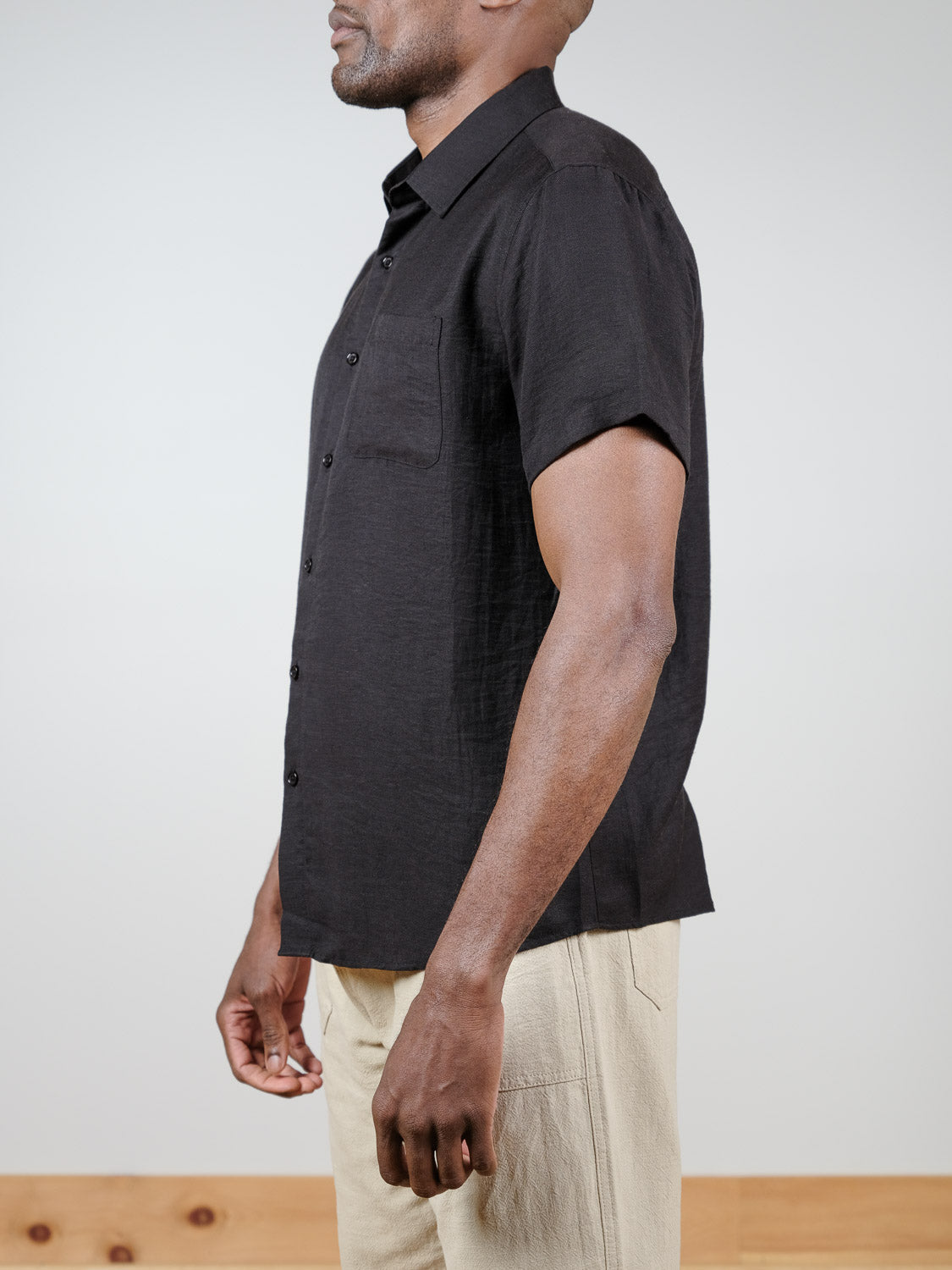 A man stands in profile wearing the Hiker Short Sleeve Shirt by ID Menswear—a relaxed-fit black button-up with a chest pocket—paired with light beige pants, highlighting effortless summer basics against a simple backdrop.