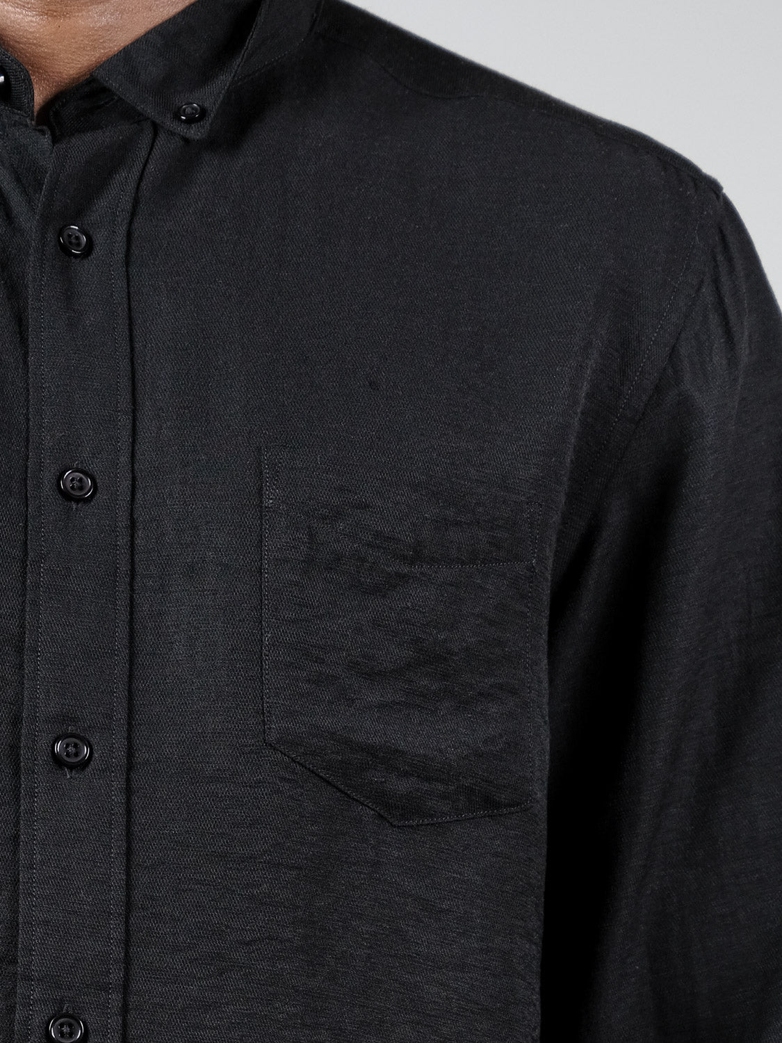 A close-up showing the upper torso and collar of a person wearing the ID Menswear Lagos Long Sleeve Shirt, a relaxed fit black button-up with a chest pocket, against a plain, light-colored background.