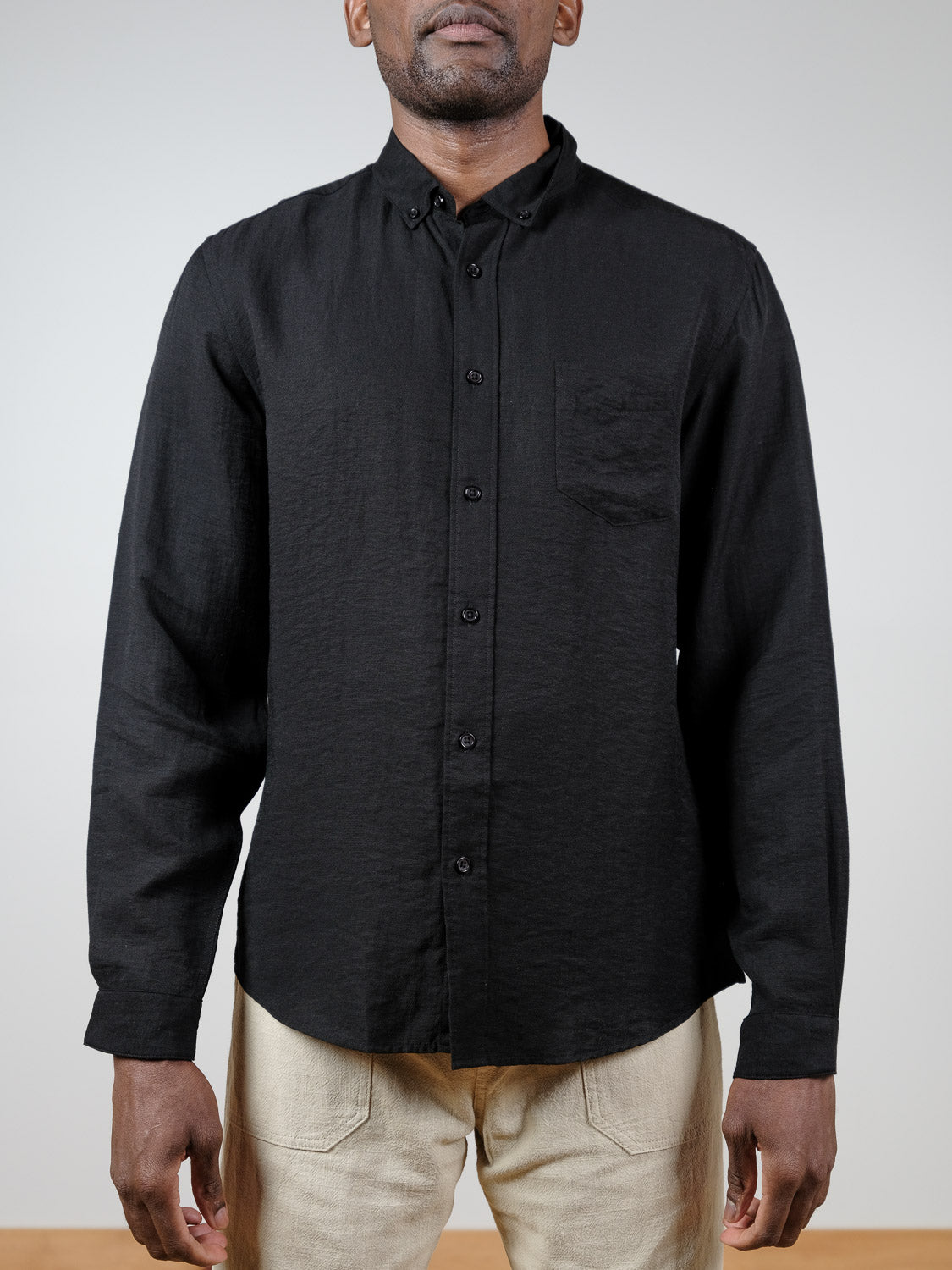 A person stands against a plain light background wearing an ID Menswear Lagos Long Sleeve Shirt in black—breathable linen with a chest pocket and relaxed fit—paired with light beige pants.