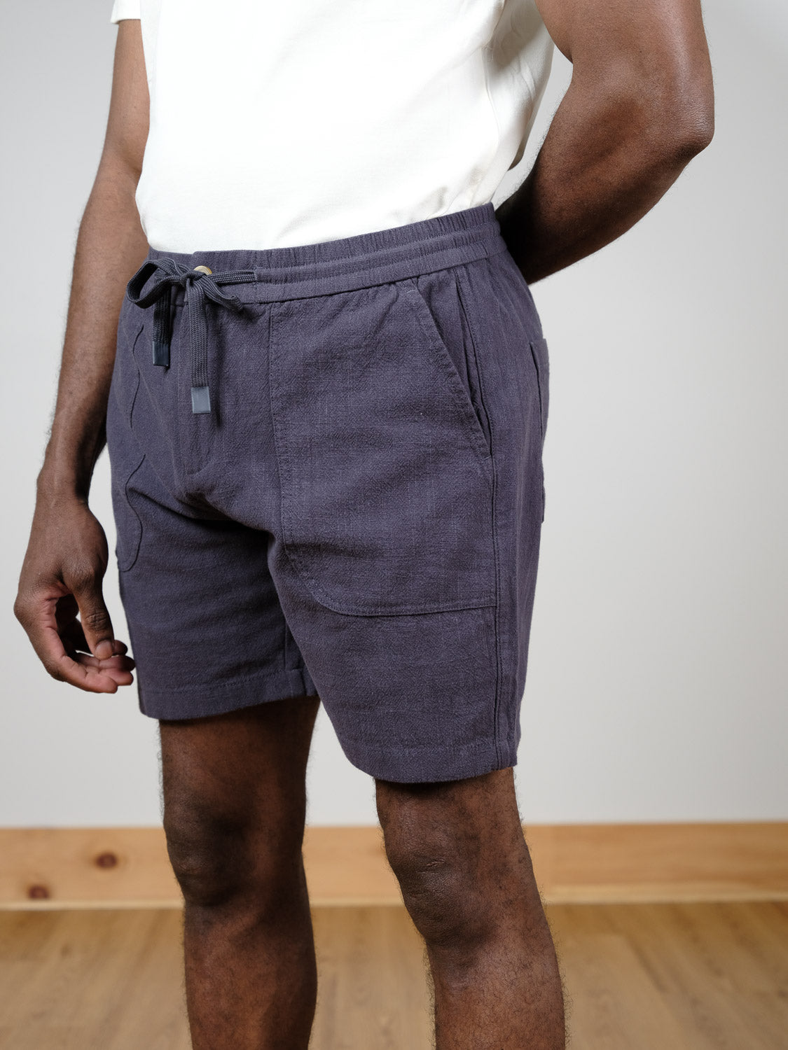 A person stands indoors on a wooden floor against a plain light wall, wearing a white shirt and dark blue Phi Phi Linen Blend Drawstring Short by ID Menswear, with one hand by their side and the other behind their back.