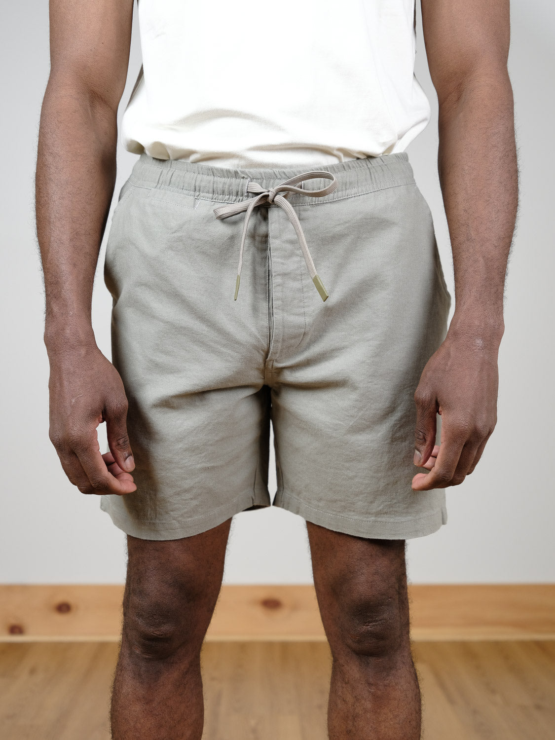 A person stands indoors on a wooden floor, wearing a white shirt and the ID Menswear Kotao Linen Blend Drawstring Short in light gray, arms relaxed at their sides.