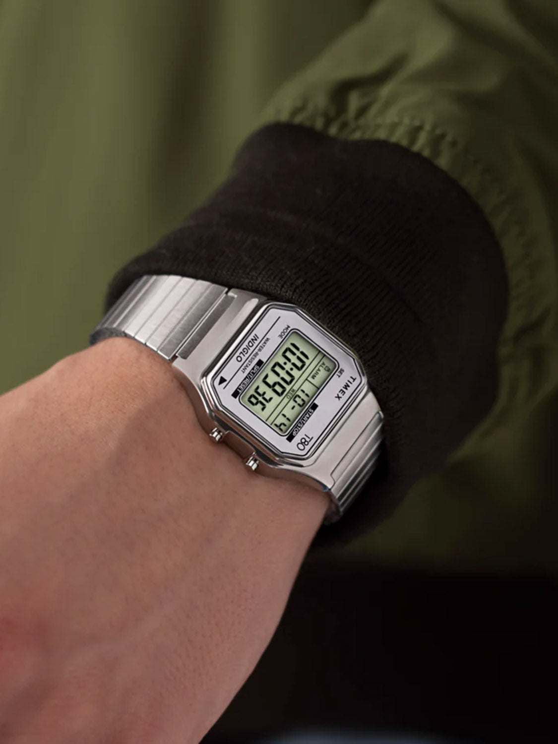 Close-up of someone wearing a Timex T80 34mm Stainless Steel Expansion Band Watch. The silver digital watch, with a retro design, shows the time as they wear a dark green and brown long-sleeve jacket.