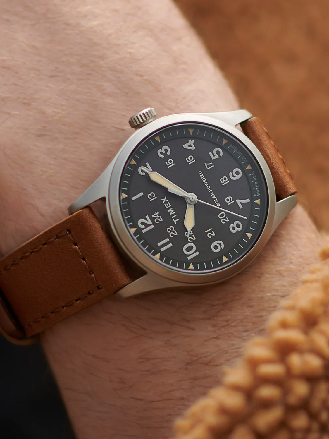 A close-up of a wrist wearing the Timex Expedition Field Post Solar 36mm watch with an eco-friendly brown leather strap and black dial with white numerals, paired with a tan textured sleeve.