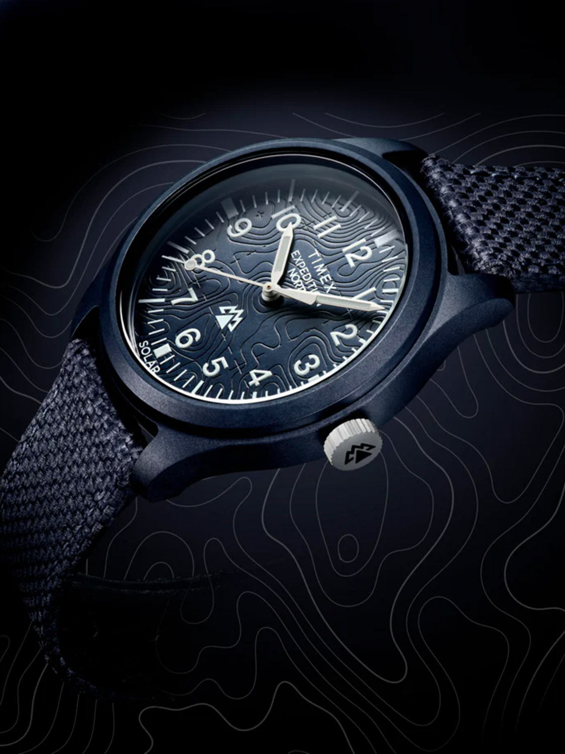 Close-up of the Timex Expedition Traprock Solar 40mm featuring a textured dial with large luminous numbers. The eco-ceramic case houses solar-powered movement, and it has an eco-friendly fabric strap against a dark, swirling backdrop.