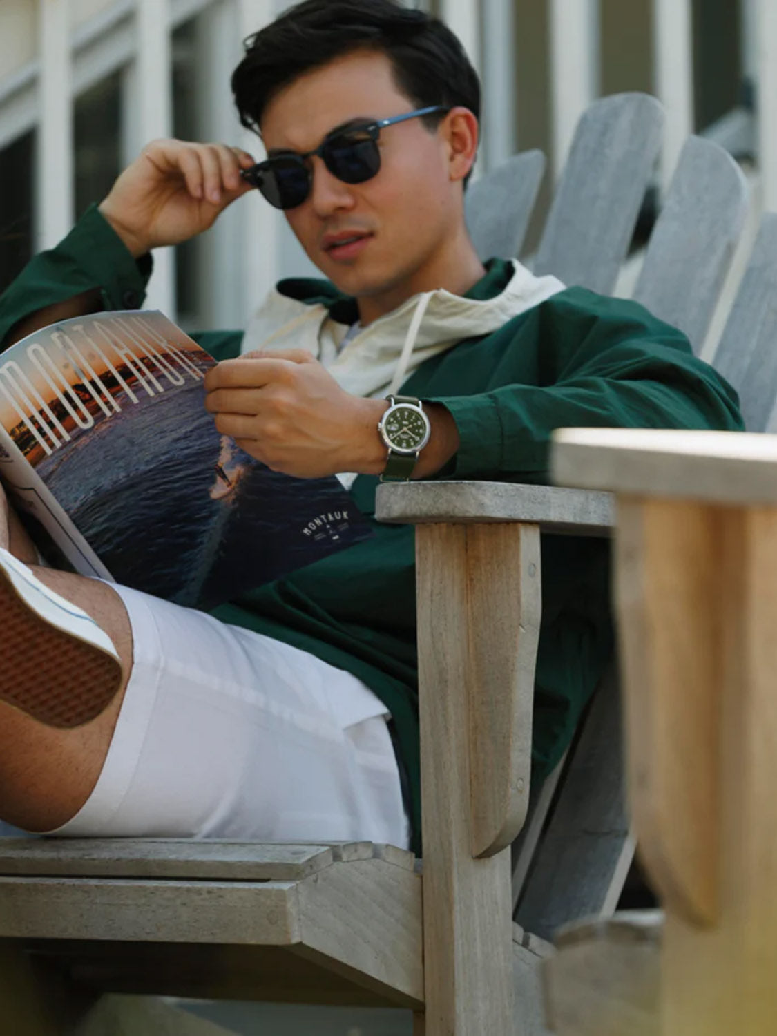 Relaxing in a wooden chair, wearing sunglasses and a green jacket, they hold a magazine. They sport white shorts and an eco-friendly Timex Weekender 40mm Leather Strap Watch featuring an INDIGLO® backlight, embodying a leisurely weekender vibe.