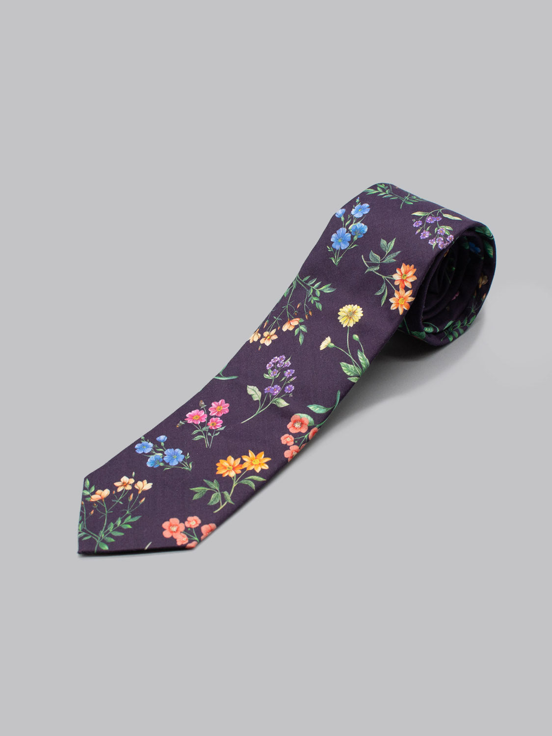 The ID Menswear Liberty of London Tana Lawn™ Annie is a handmade, dark purple necktie with a colorful floral pattern in red, yellow, blue, and orange, displayed rolled up on a plain gray background.
