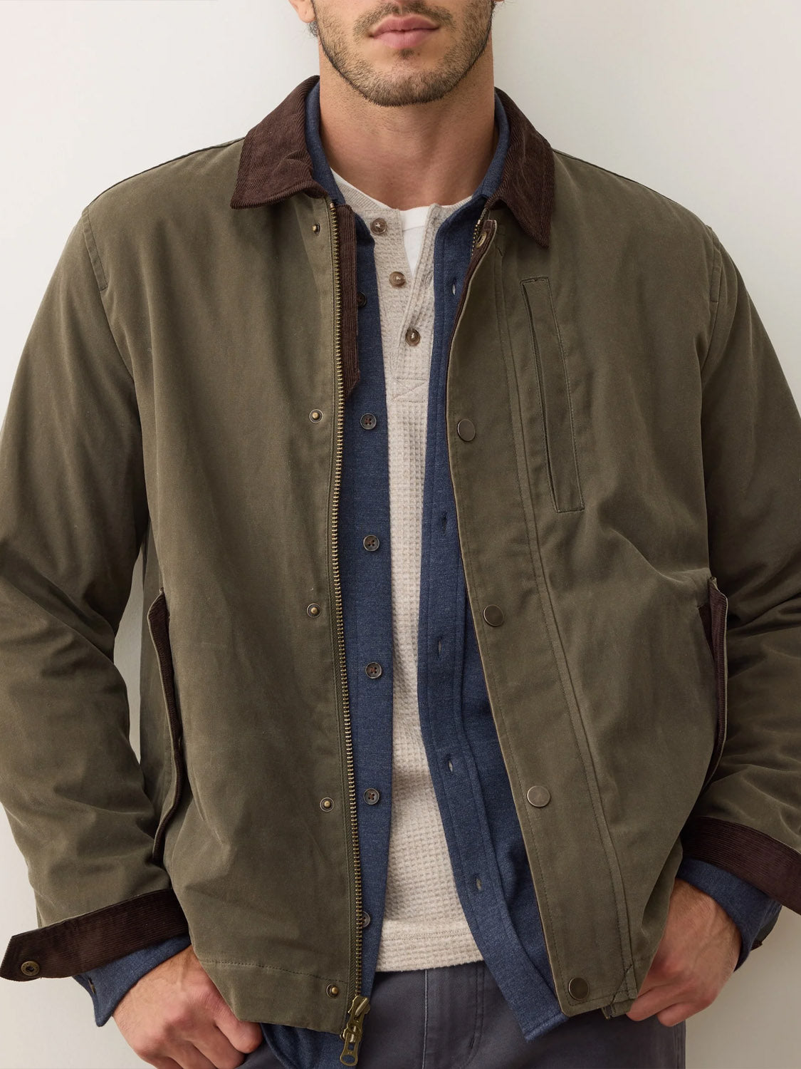 A man wears the Marine Layer Weston Wax Canvas Barn Jacket with a brown collar, layered over a blue button-up and a white waffle-knit henley, hands in pockets, face partially out of frame.