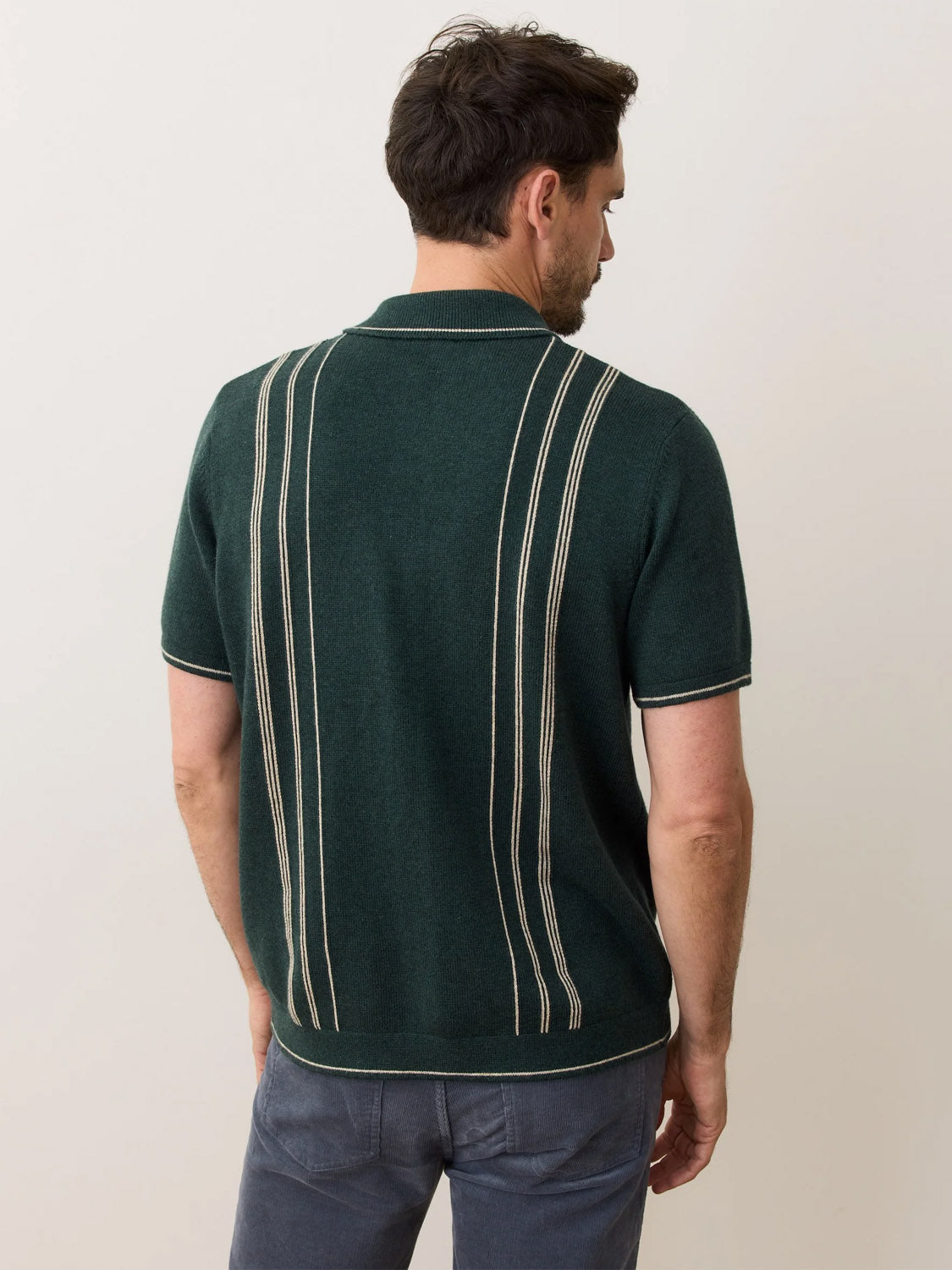 A man with short dark hair wears Marine Layer's Barry Sweater Button Down, a classic fit dark green knit shirt with cream vertical stripes and a collar, paired with dark gray pants against a plain light background.