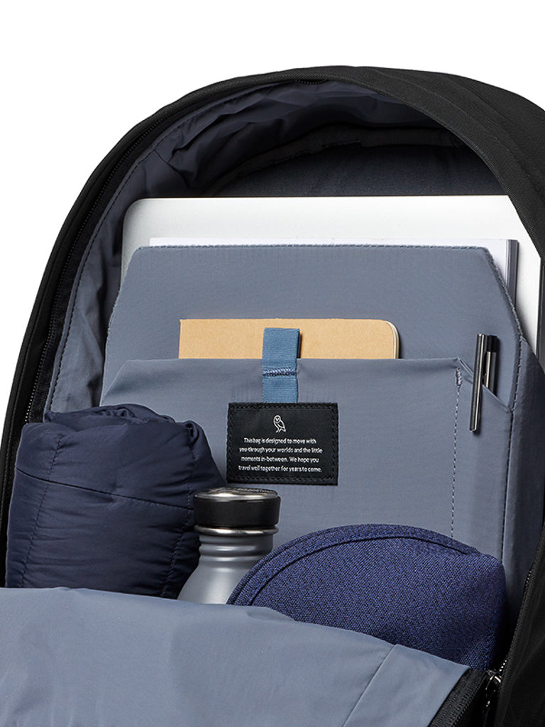 The Bellroy Classic Backpack - 20L, made from recycled materials, displays a laptop, notebook, pen, jacket, reusable water bottle, and a textured blue fabric item, all neatly organized in separate compartments.