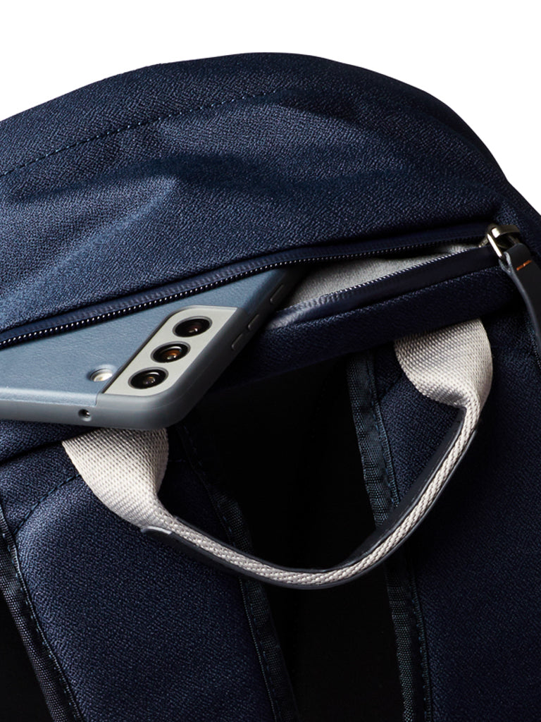 A close-up of the Bellroy Classic Backpack - 20L in navy blue, made from recycled materials, shows a gray smartphone partly in a zippered pocket with visible camera lenses. The backpack features white fabric handles.