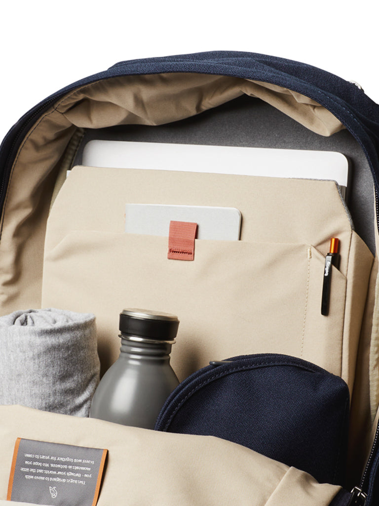 The Bellroy Classic Backpack - 20L, made from recycled materials, is open to reveal a laptop, notebook with red tab, pen, water bottle, grey rolled shirt, and navy pouch neatly stored in beige interior pockets.