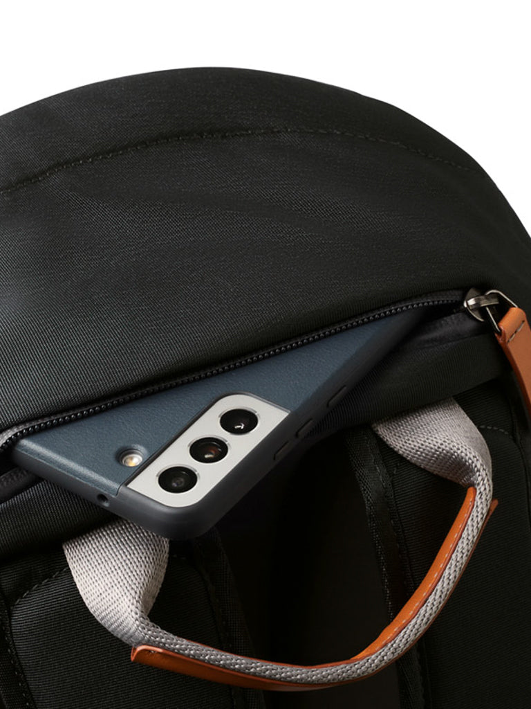 A close-up of the Bellroy Classic Backpack - 20L shows its zippered pocket open with a smartphone inside. The pack features gray and brown straps, smooth, durable recycled fabric, and sustainable craftsmanship.