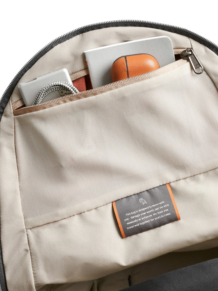 A close-up of the beige Bellroy Classic Backpack - 20L shows its zippered pocket storing a laptop, orange mouse, charging cable, and book. Made from recycled materials, it features a small tag with text sewn below the pocket.