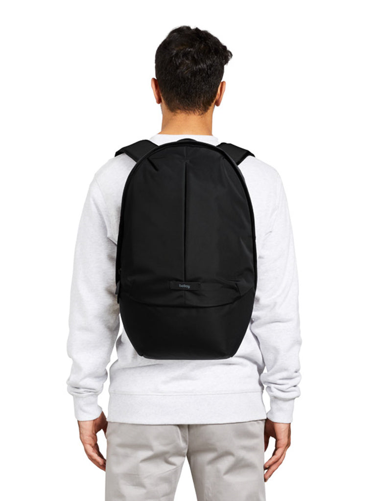 A person wearing a white sweatshirt and light gray pants is seen from behind, carrying the Bellroy Classic Backpack Plus - 24L, a large black backpack with dual compartments and padded shoulder straps.