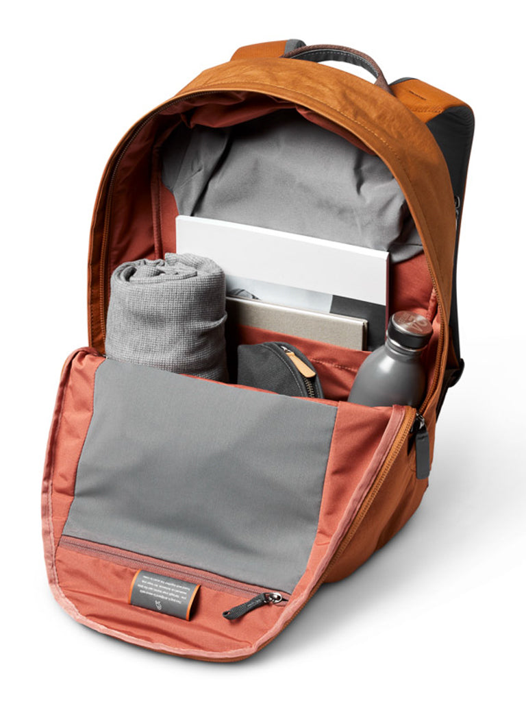 The open Bellroy Classic Backpack Plus - 24L, in amber, reveals a rolled-up gray sweatshirt, laptop, notebook, water bottle, and zip pouch inside organized pockets made from water-resistant recycled fabric.