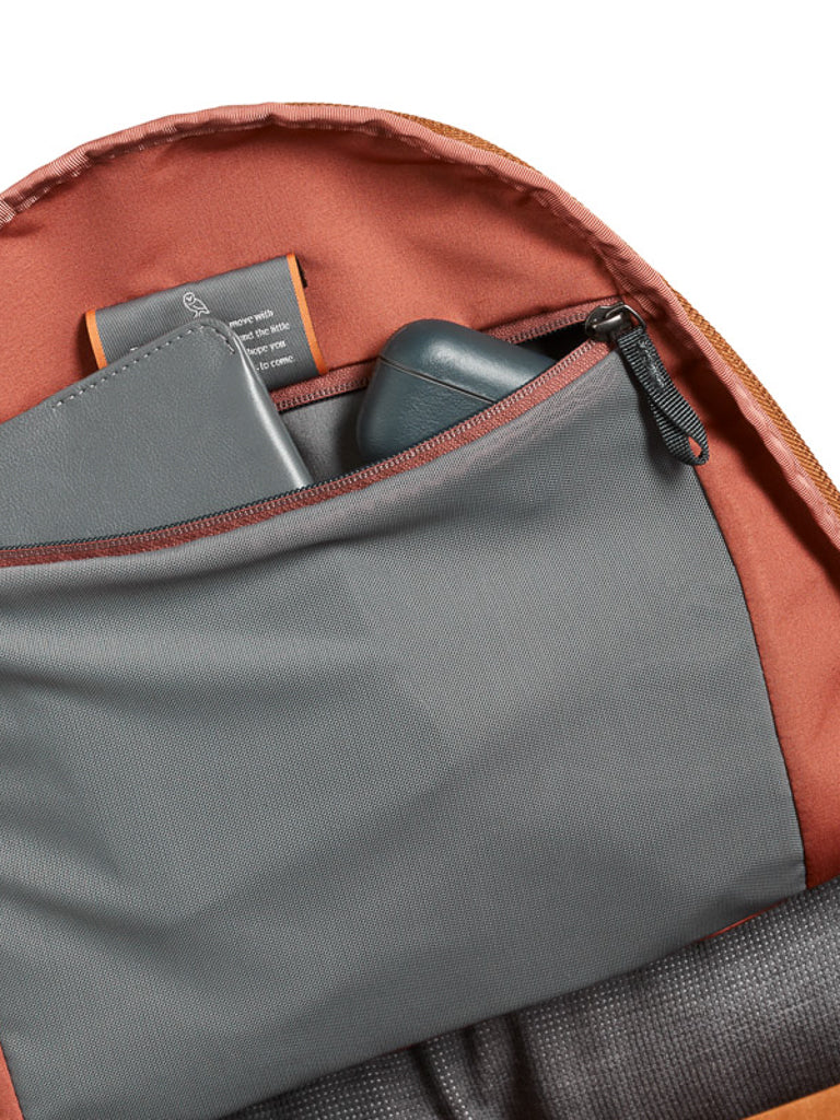 The Bellroy Classic Backpack Plus - 24L features a dual compartment with a gray zippered pocket, storing a gray notebook and dark wireless earbuds. Its reddish-brown lining complements the water-resistant recycled fabric exterior.