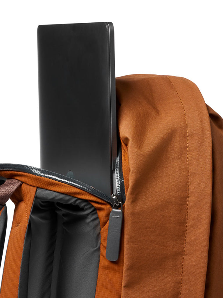 A close-up of the Bellroy Classic Backpack Plus - 24L in brown, featuring dual compartments and a partially unzipped section with a black laptop being placed inside.