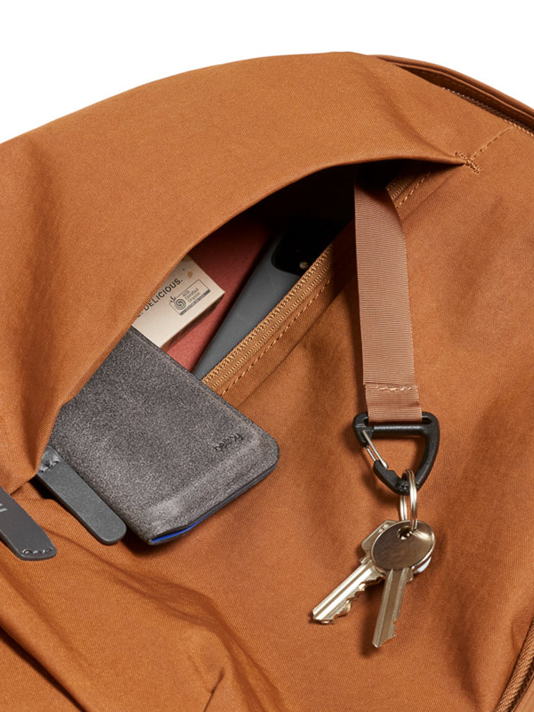 A close-up of Bellroy's Classic Backpack Plus - 24L, crafted from water-resistant recycled fabric, shows its open dual compartments holding a gray wallet, smartphone, red item, and two silver keys on a keyring attached to a strap.