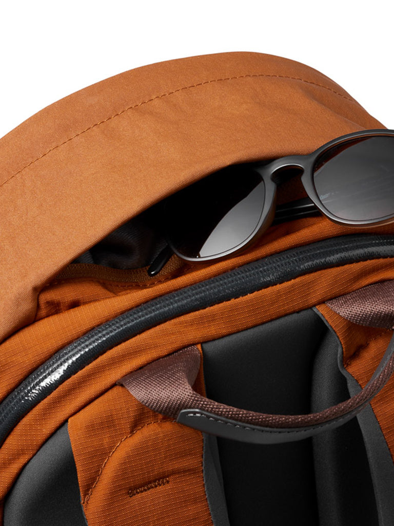 A close-up of the Bellroy Classic Backpack Plus - 24L in orange shows its dual compartments, partially unzipped front pocket with black sunglasses inside, brown straps, and water-resistant recycled fabric.