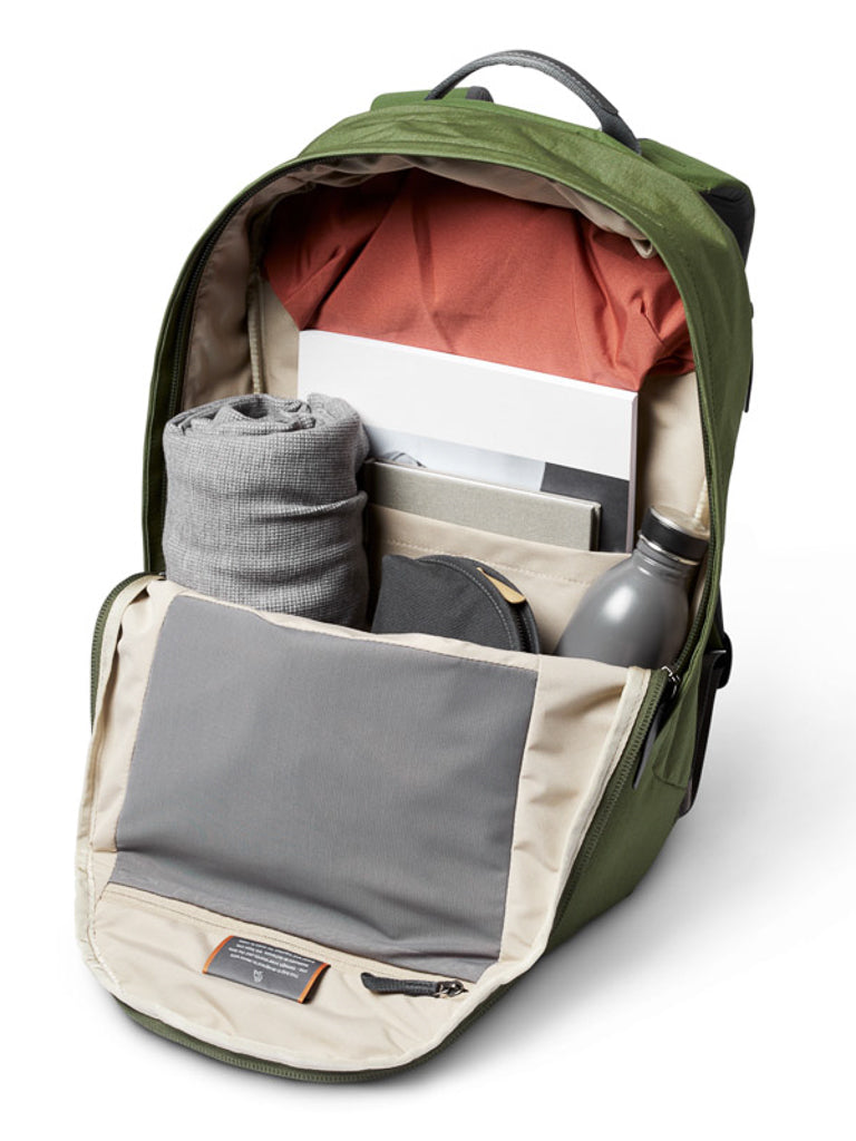 The Bellroy Classic Backpack Plus - 24L, made from water-resistant recycled fabric, features dual compartments and opens to reveal a red shirt, rolled gray garment, water bottle, laptop, books, and a small zippered pouch—neatly organized inside.