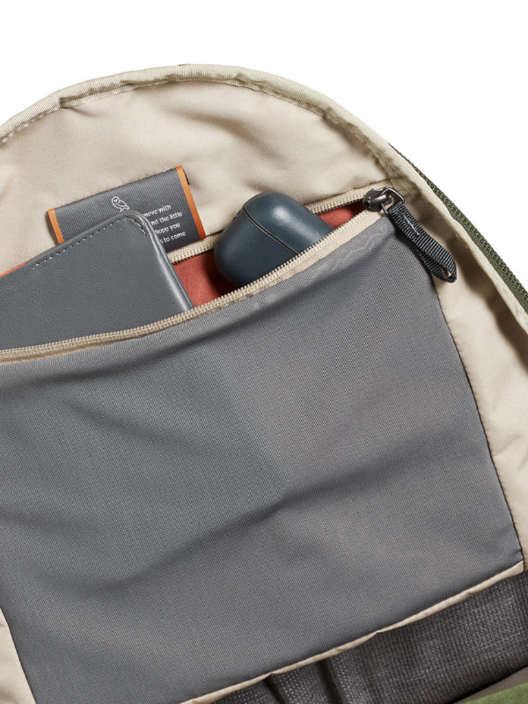 A close-up of the Bellroy Classic Backpack Plus - 24L, made from water-resistant recycled fabric, shows a beige-lined gray zippered pocket holding a gray notebook, dark wireless earbud case, and a small orange-bound book.