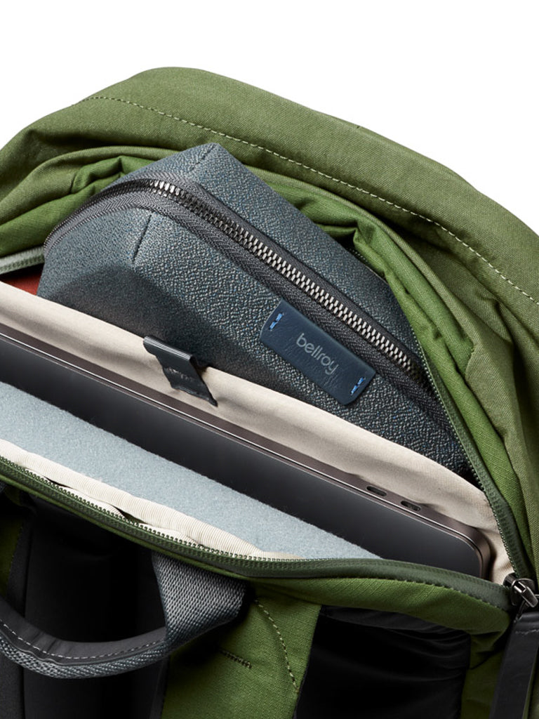 A Bellroy Classic Backpack Plus - 24L in green recycled fabric is shown open, with a gray Bellroy zip pouch, closed laptop, and cream folder neatly organized inside.