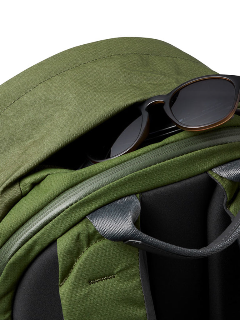 A pair of black sunglasses is partially tucked into the top pocket of a green Bellroy Classic Backpack Plus - 24L, made from water-resistant recycled fabric, with visible shoulder straps in the foreground.