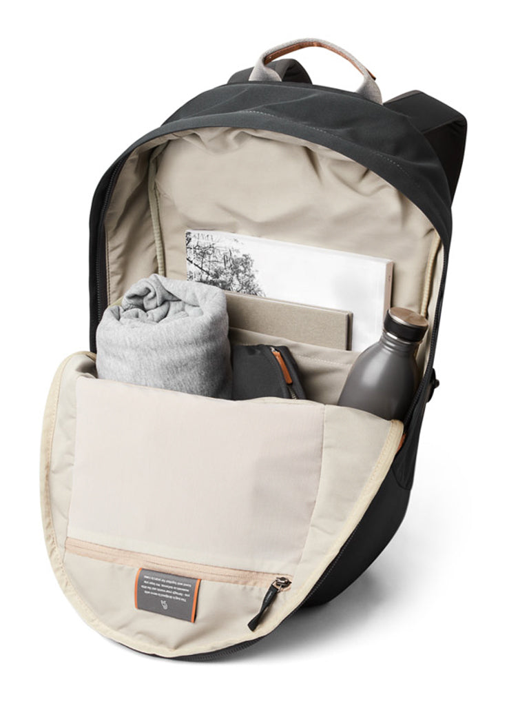 The Bellroy Classic Backpack Plus - 24L is a black, dual compartment pack made from water-resistant recycled fabric, shown with its main section open to reveal a hoodie, stainless steel water bottle, notebook, illustrated book, and small pouch inside.